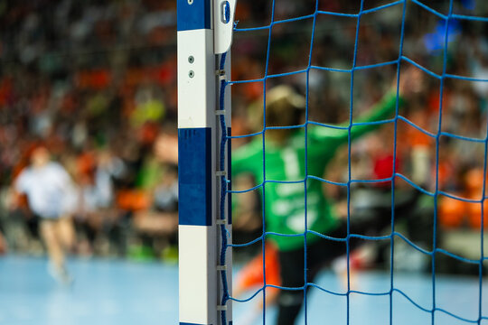 Handball goalkeeper defending the goal during a professional indoor match, viewed through the goal net with intense game atmosphere