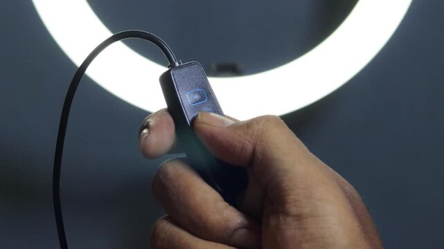 Hand of an Asian man adjusting the brightness of a glowing LED ring light using an inline controller, with illuminated control buttons against a dark background.