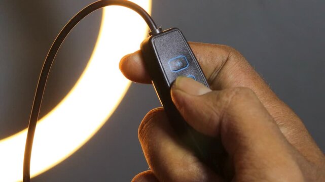 Hand of an Asian man adjusting the brightness of a glowing LED ring light using an inline controller, with illuminated control buttons against a dark background.