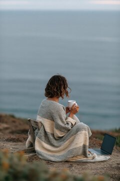 Woman wrapped in striped blanket sitting on a low chair by the ocean holding a mug with laptop nearby for cozy morning background and remote work banner