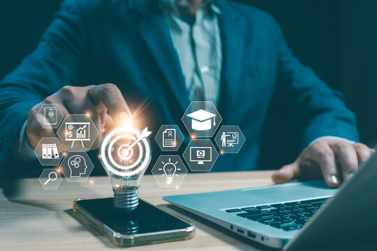 Businessman touching light bulb with target goal icon, digital education graphics on smartphone and laptop, concepts of smart learning innovation, knowledge and business achievement.