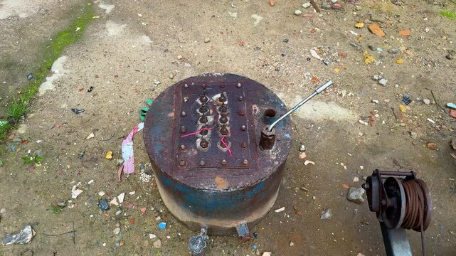 Rusty industrial metal cylinder with electrical terminals and lever standing outdoors beside a cable winch, old equipment, corrosion, industrial machinery
