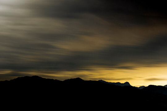 Dramatic clouds and silhouettes of mountains at sunset. A captivating long exposure image showcasing dark clouds sweeping over a mountain silhouette, creating a moody and atmospheric landscape.