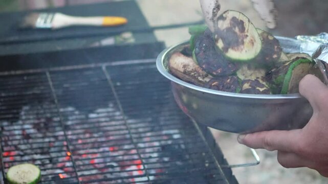 Healthy picnic catering with roasted zucchini rounds in a stainless steel bowl near hot coals. Static close-up shot.