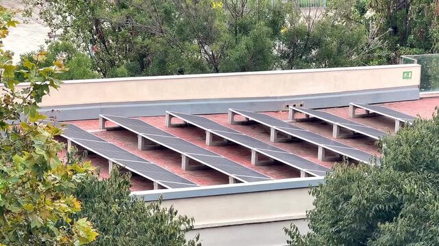 High angle shot of a row of photovoltaic panels on a building roof among green trees, generating clean green energy. Barcelona Spain 16,09,2026