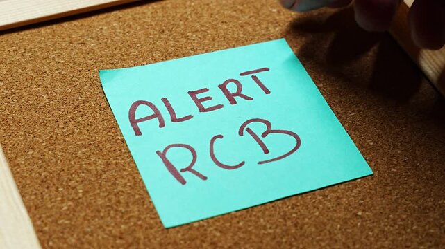 Handwritten ALERT RCB on a turquoise memo fills the frame over brown cork, with the board edge in the corner. It represents government SMS warnings, phone emergency messages, and flood and storm aler