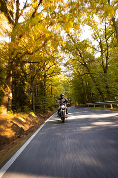 A motorcyclist overtakes a small yellow car on an asphalt road through the forest. The moving shot captures both vehicles in motion against a wooded roadside backdrop.