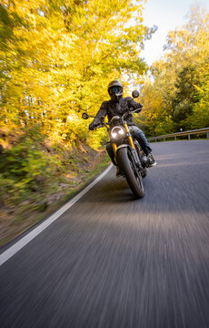 A motorcyclist overtakes a small yellow car on an asphalt road through the forest. The moving shot captures both vehicles in motion against a wooded roadside backdrop.