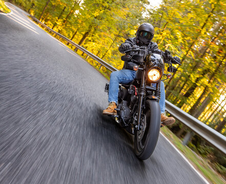 A motorcyclist overtakes a small yellow car on an asphalt road through the forest. The moving shot captures both vehicles in motion against a wooded roadside backdrop.