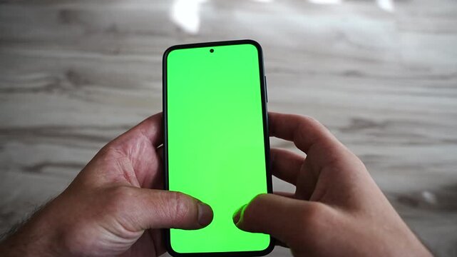 A finger taps and swipes a green screen smartphone held in the other hand, seen from a first person angle above a wooden table. A touchscreen chroma key for app navigation, online ordering, booking a