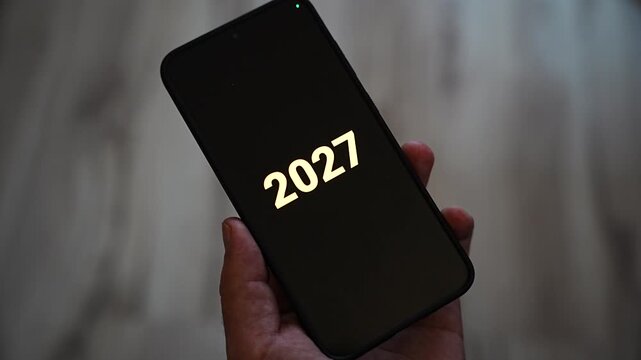 Golden digits 2027 appear one after another on a black smartphone screen held upright in one hand. A clean year number for upcoming year announcements, annual goals, new year countdown and calendar 2