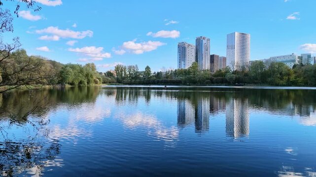Calm city lake reflects modern high rise buildings and white clouds under a blue sky.