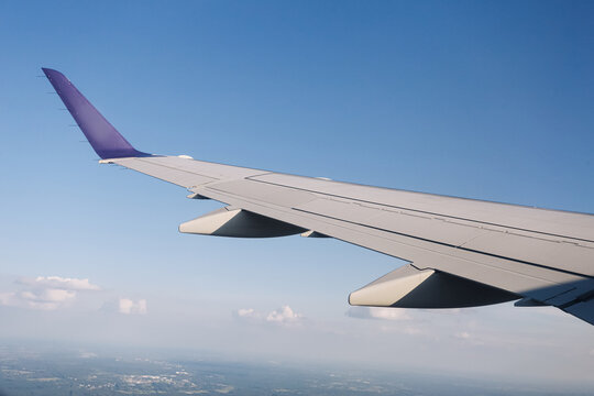 Passenger airplane wing above landscape. Purple wingtip against blue sky. White clouds near distant horizon. Aerial countryside beneath aircraft wing. Daylight flight travel background.