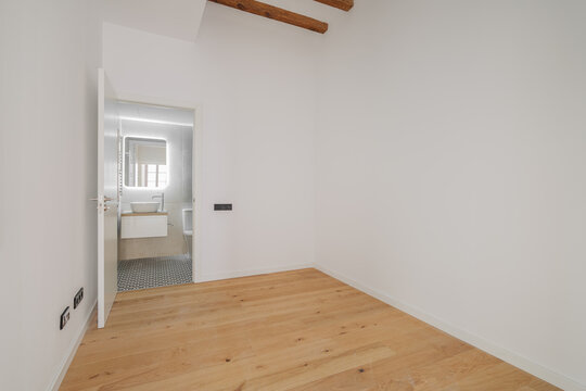 A modern, empty room with white walls and wooden flooring. A doorway leads to a bathroom area. Natural light enters through a window.