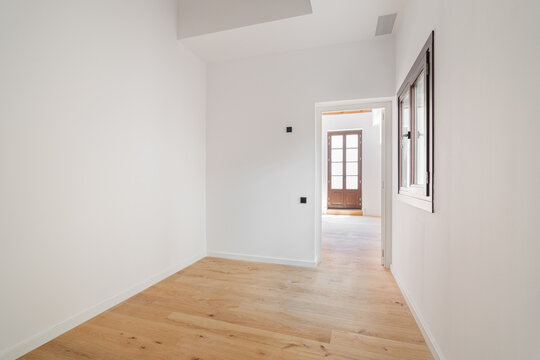 Empty room with white walls and wooden floor. A doorway leads to another room with natural light. Minimalist design and spacious layout.
