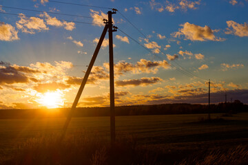 Fototapeta premium Utility poles silhouette against a vibrant October sunset over open autumn fields, highlighting electrical power lines crossing the rural landscape during golden hour