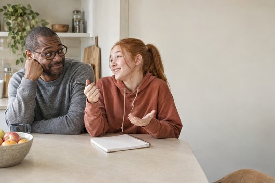 Stepfather and teenage stepdaughter negotiating pocket money with a notepad at the kitchen table, blended family finances with copy space