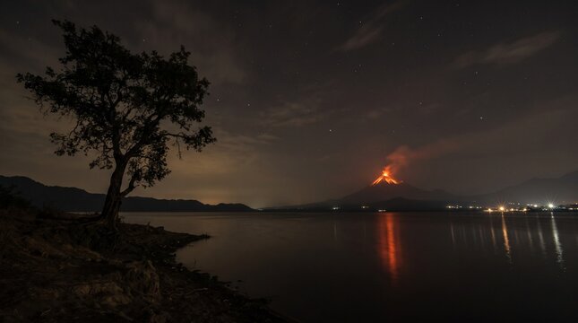A dramatic night view of an erupting volcano reflecting in a calm lake with a lone tree silhouette in the foreground.