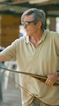Concentrated senior man with sunglasses practicing traditional archery. An elder archer holding a wooden longbow and preparing to shoot. Vertical
