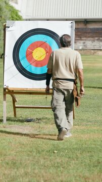 Senior man in sportswear walking towards a target to retrieve his arrows. Outdoor archery practice on a sunny day in a green field. Vertical