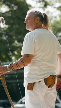 Senior man with a ponytail practicing archery. Focused archer aiming and shooting with a wooden longbow in a sunlit forest environment. Vertical