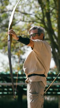 Senior male archer wearing sunglasses concentrating on aiming his longbow. This athlete is practicing his favorite sport on a beautiful sunny day. Vertical