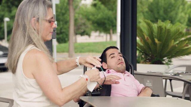 Caring woman cleaning and putting on glasses for a young man with spina bifida sitting in a wheelchair. Quality time and assistance concept