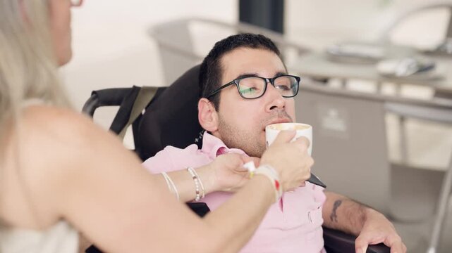 Caring woman assisting a young man with spina bifida sitting in a wheelchair. She is helping him drink a cappuccino from a mug