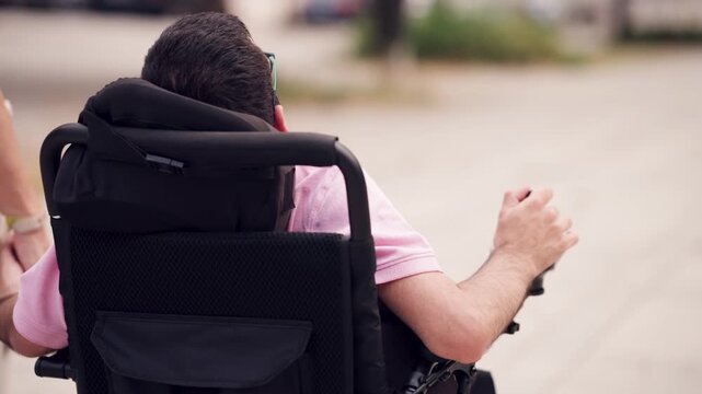 Rear view of a young man with spina bifida navigating an electric wheelchair outdoors. His hand is controlling the joystick for mobility