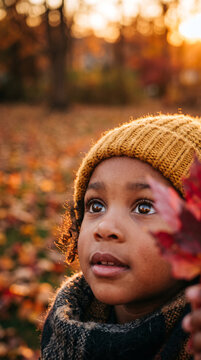 Warm autumn light bathes a child in a knitted hat. Child gazes upward, eyes wide with wonder. Scarf wraps snugly around their neck, cozy and soft. Leaves carpet the ground, golden and red
