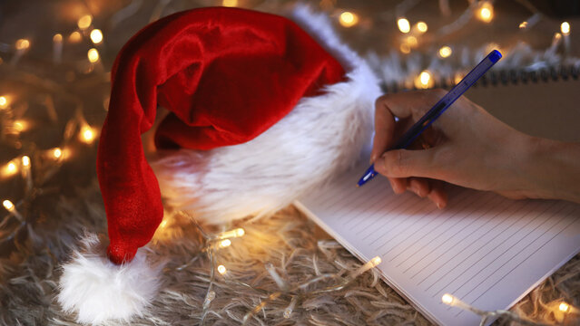 Hand writing beside Santa hat, holiday stationery and Christmas gift planning with wish lists and letters to Santa