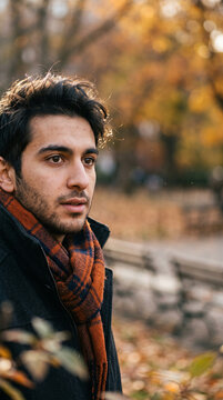 Young man gazes thoughtfully at autumn scene. Golden leaves frame his warm, contemplative expression. Cozy scarf and coat suggest crisp fall air. He stands near park bench. Perfect for lifestyle