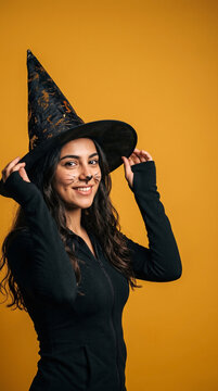 Woman smiles warmly, adjusting her black witch hat. Long curls frame her face as she looks toward the camera. She wears a black top against a solid orange background