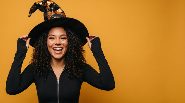 Woman wears witch hat, smiles brightly. Curly hair frames her cheerful face. Hands adjust hat as she poses. Bright orange background, clean, bold. Perfect for Halloween or fantasy themes