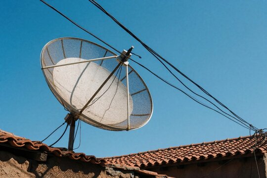 Large mesh satellite dish antenna installed on rooftop of house. White parabolic receiver connects to electrical wires against clear blue sky. Telecom equipment provides media signal, internet