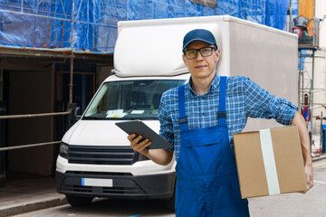 A delivery driver holding a cardboard box and a tablet in front of a white cargo van. © scharfsinn86
