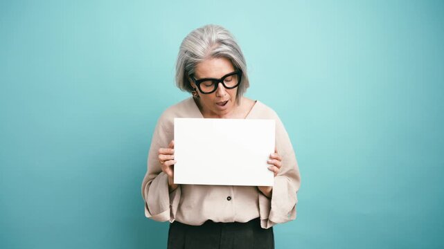 Expressive senior woman wearing eyeglasses holding a white blank board for text or advertising. Isolated on a plain blue background