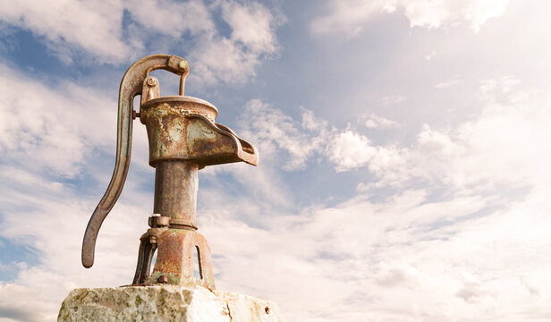 An old rusty hand water pump against a cloudy atmospheric background symbolizing water scarcity and drought.