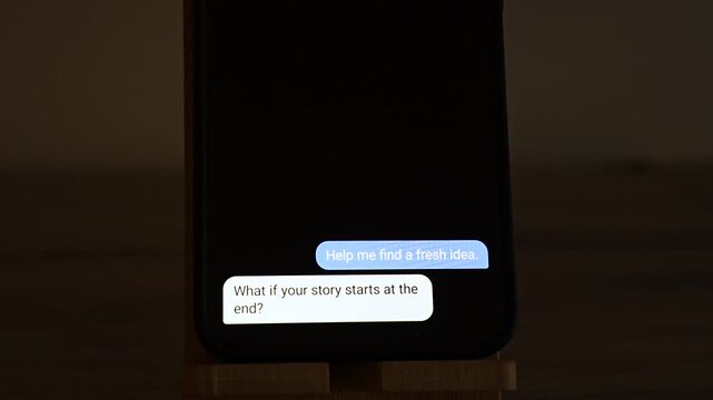 Chat bubbles on an upright smartphone show a user asking for a fresh idea, an AI suggesting a story that starts at the end and a happy reply. A picture of AI brainstorming, beating writer block and c