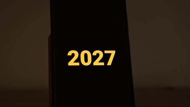 Big golden yellow 2027 numerals sit centered on a black smartphone screen standing in a warm, dim room. A simple new year concept for the upcoming year, annual planning, calendar headers and 2027 ann