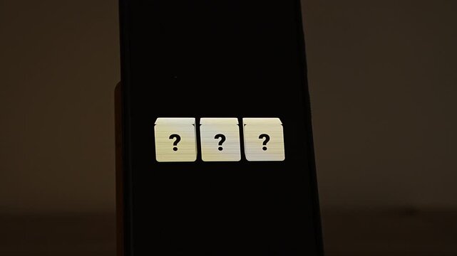 Three cream cubes with black question marks line up on a smartphone standing in a dark, warm room. A visual for questions, uncertainty, mystery, FAQ, quiz time and unknown answers waiting to be revea