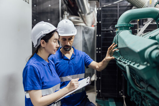 Two engineers in hard hats inspect a large industrial generator, reviewing notes on a clipboard during maintenance inside a mechanical room.