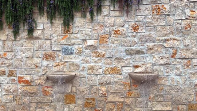 Two Decorative Wall Fountains with Flowing Water on Stone Wall. Water flowing from two decorative wall fountains against a textured stone wall with hanging greenery. Peaceful garden design.