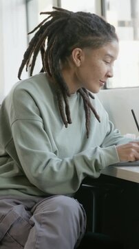 Vertical video: Drawing one with locs in green top with pen on tablet at window, light causing rest
