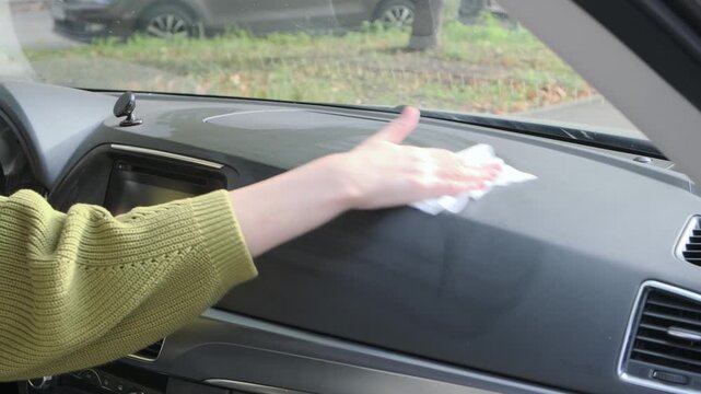 Focused female driver cleans front dashboard surface inside car with wet wipe, removing accumulated dust particles across dark plastic panel during thorough interior maintenance routine