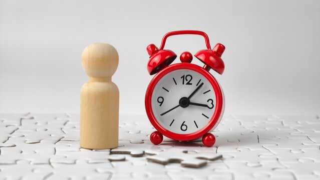 Time management and problem solving concept with wooden peg doll and red alarm clock standing on jigsaw puzzle

