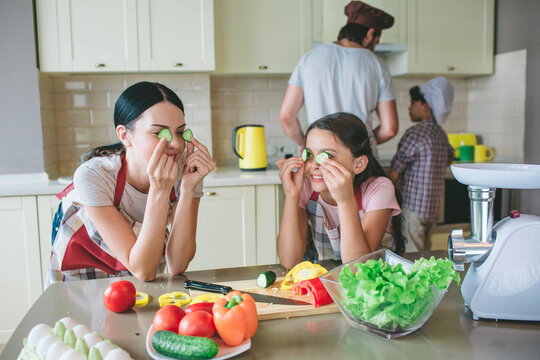 Moom and daughter stands together and leans at table. They are playing with round pieces of cucumber. They put it on eyes. Boys are working at stove. They cook breakfast.