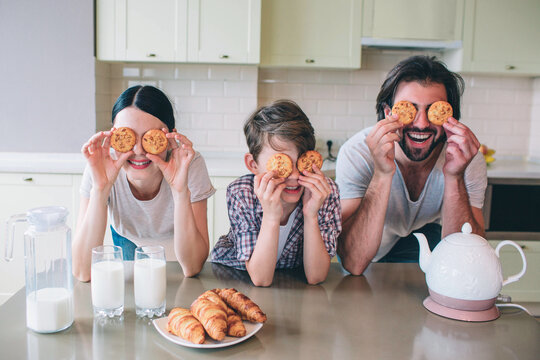 Happy family are playing with rolls. They pyt them on eyes and smiling. Family are leaning to table. There are kettle, milk and croissans on it.