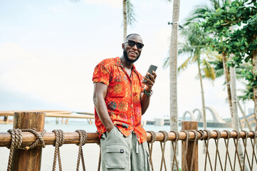 A man stands on a sunny tropical beach checking his smartphone while wearing a red patterned shirt and sunglasses. Casual vacation vibes and modern lifestyle combine as he texts or scrolls near a mari © Lumos sp