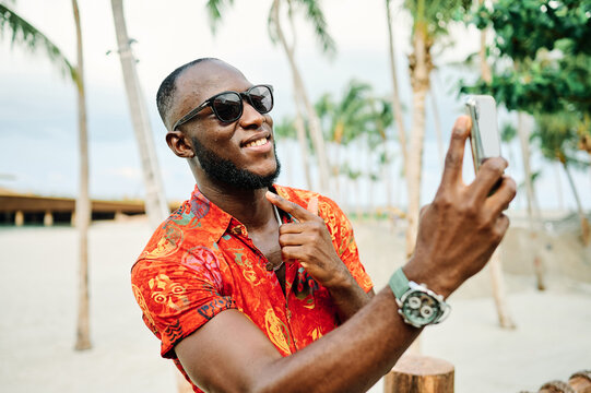 A smiling man takes a selfie on a tropical beach lined with palm trees while wearing sunglasses and a bright patterned shirt. He holds a smartphone, enjoying a relaxed vacation moment at a sunny resor